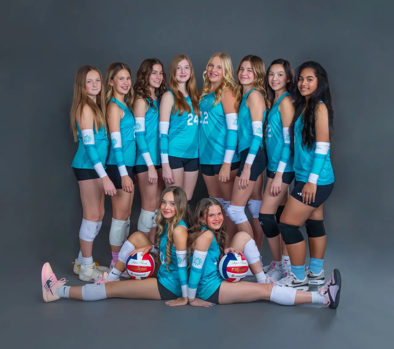 Volleyball team in teal uniforms posing in studio, with two players seated in front holding volleyballs