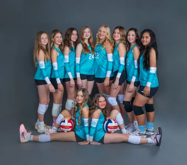 Volleyball team in teal uniforms posing in studio, with two players seated in front holding volleyballs